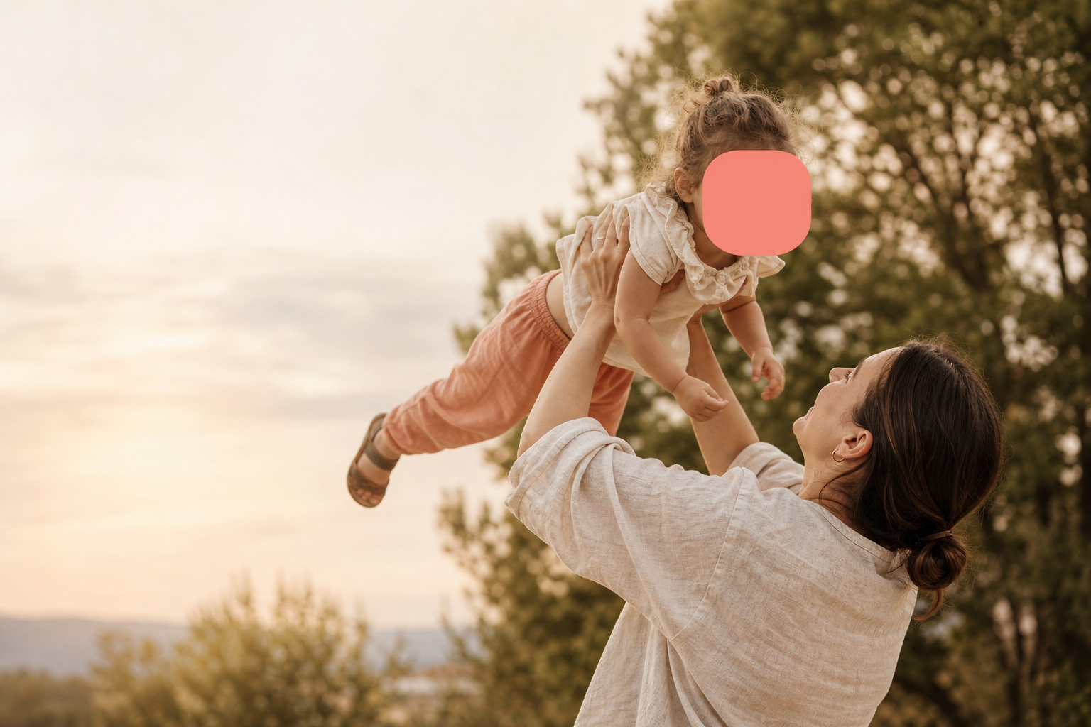 A parent lifting their toddler into the golden-hour light, the child's face gently covered by a soft coral block